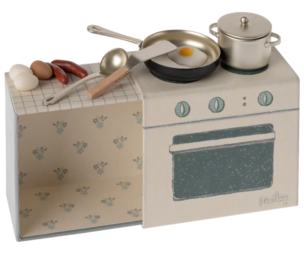 A Maileg Cooking Set designed for mice, featuring a miniature toy stove with a frying pan containing a sunny-side-up egg, a pot, sausages, and three eggs on the countertop. This small kitchen set includes kitchen utensils and an oven, all set against a floral-patterned background—perfect for any kids playset.