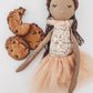 The MON AMI Cookie Scented Doll by Mon Ami, featuring an embroidered face and wearing a floral dress with a tulle skirt, is placed beside three chocolate chip cookies on a white background.