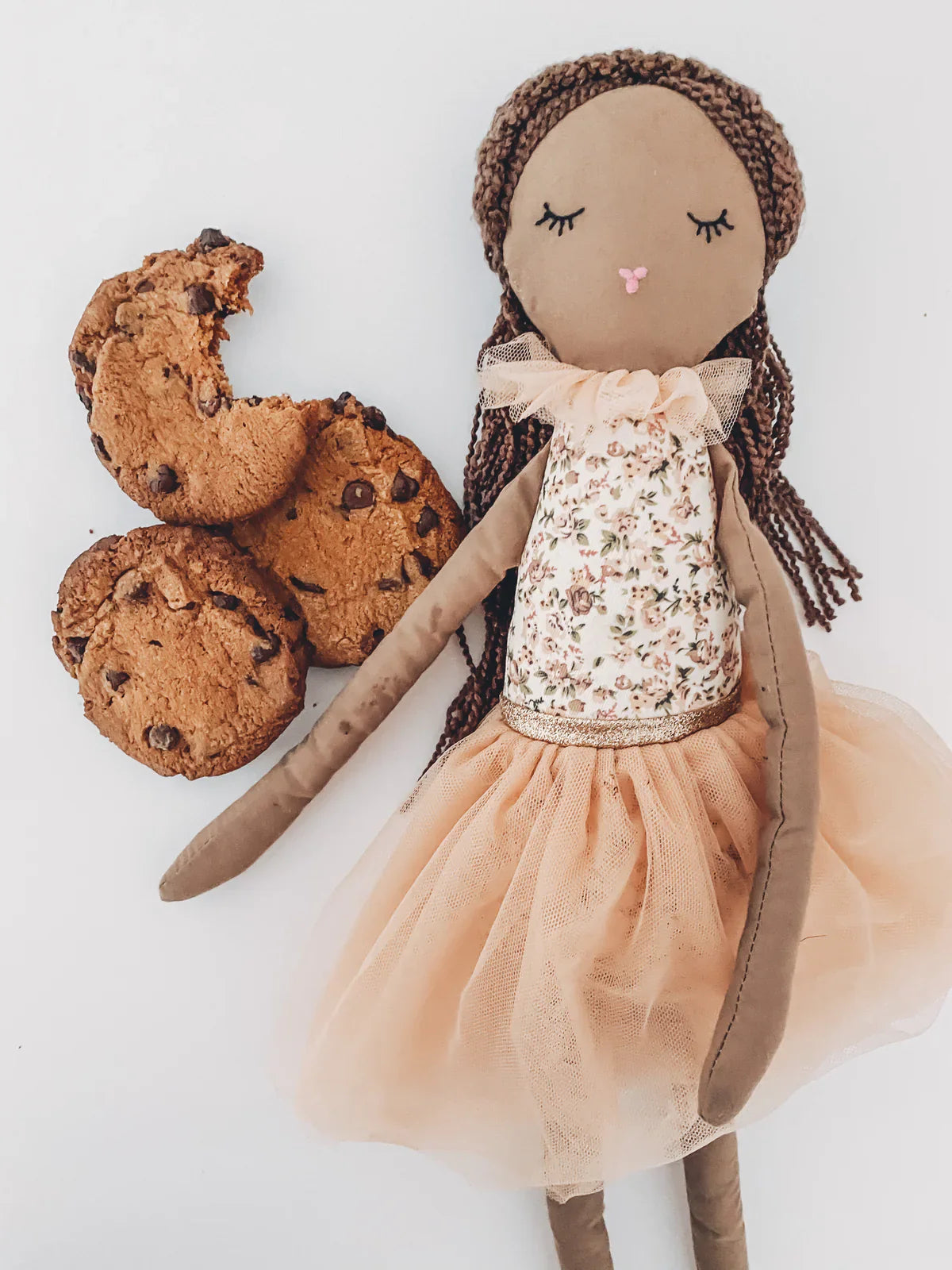 The MON AMI Cookie Scented Doll by Mon Ami, featuring an embroidered face and wearing a floral dress with a tulle skirt, is placed beside three chocolate chip cookies on a white background.