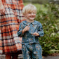 A smiling blond child wears the Pink Chicken Baby Girls’ Dawson 2-Piece Set outdoors, standing before an adult in plaid with greenery behind.