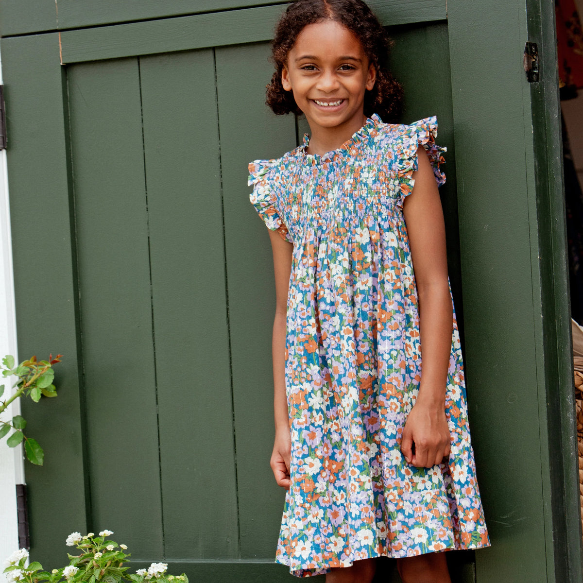 A smiling child with curly hair stands by a green door, wearing the Pink Chicken Girls’ Stevie Dress featuring ruffled sleeves and a smocked chest.