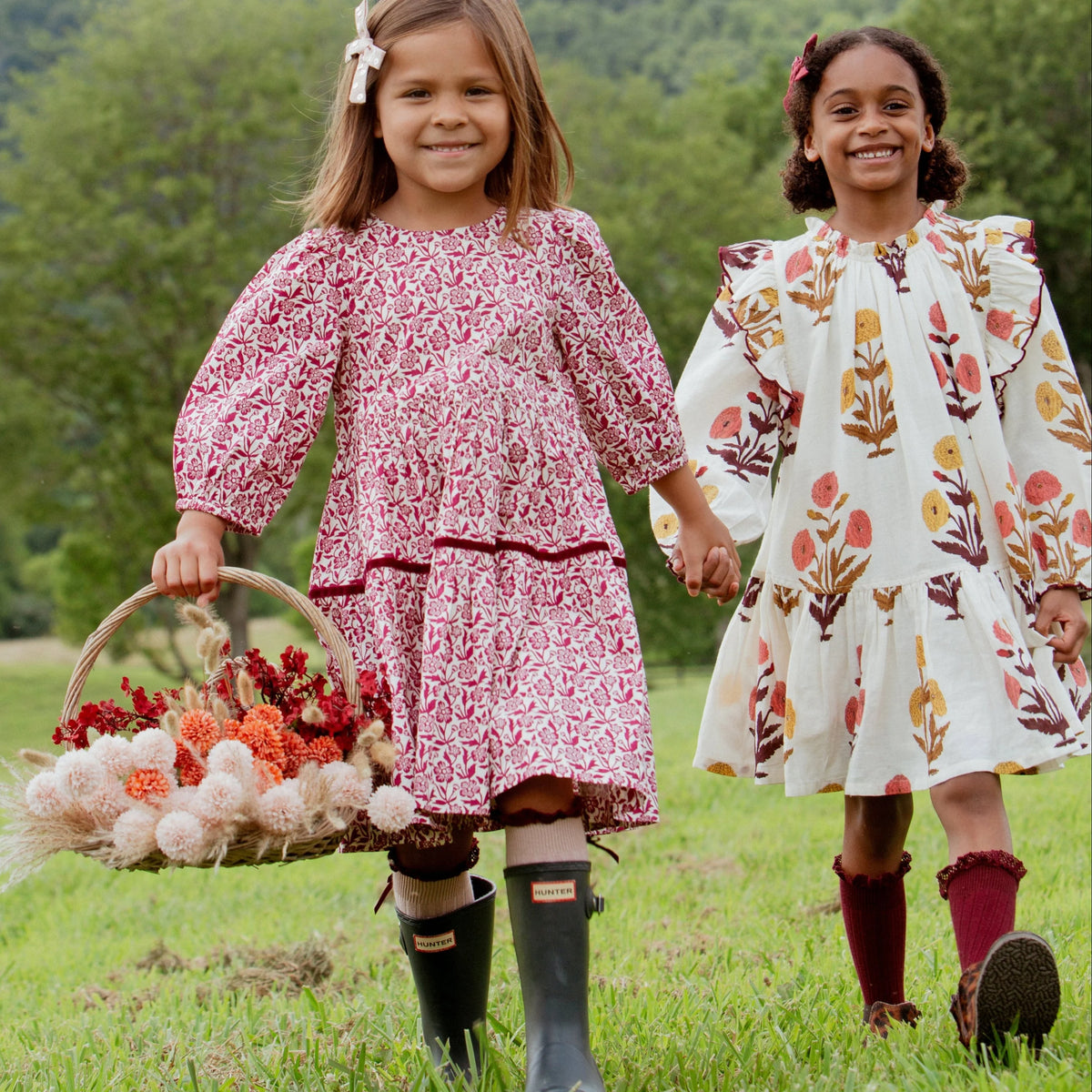 Two young girls wear the Pink Chicken Girls’ Harper Dress in Harvest Dandelion print, holding hands and a flower basket, with grassy hills and trees in the background.