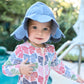 A toddler in a Pink Chicken Baby Scallop Sun Hat and a colorful shirt stands outdoors by a swimming pool.