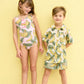 Two children stand in front of a yellow wall, wearing matching Green Banana Leaves prints; the girl wears a swimsuit, and the boy sports Pink Chicken Boys' Swim Trunks with an elastic waistband.