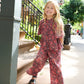 A young child with long blond hair walks along a sunny sidewalk, smiling at the camera while wearing the Pink Chicken Girls’ Jaipur Top featuring red and black floral prints, stylish bell sleeves, and navy glitter buttons, paired with black sandals.
