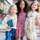 Three young girls stand together outside near a chain-link fence, two wearing crayon-patterned dresses and one in the Pink Chicken Girls’ Jennifer Dress featuring a vibrant red paisley vine pattern. One girl holds a closed book, while their colorful outfits are complemented by the tiered skirt details of the Jennifer Dress, adding playful charm to the scene.