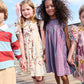 Four children stand holding hands outdoors, dressed in colorful clothes; two girls are in the center, one wearing a patterned cream Girls’ Stevie Dress by Pink Chicken and the other in a vibrant red & blue check plaid dress.