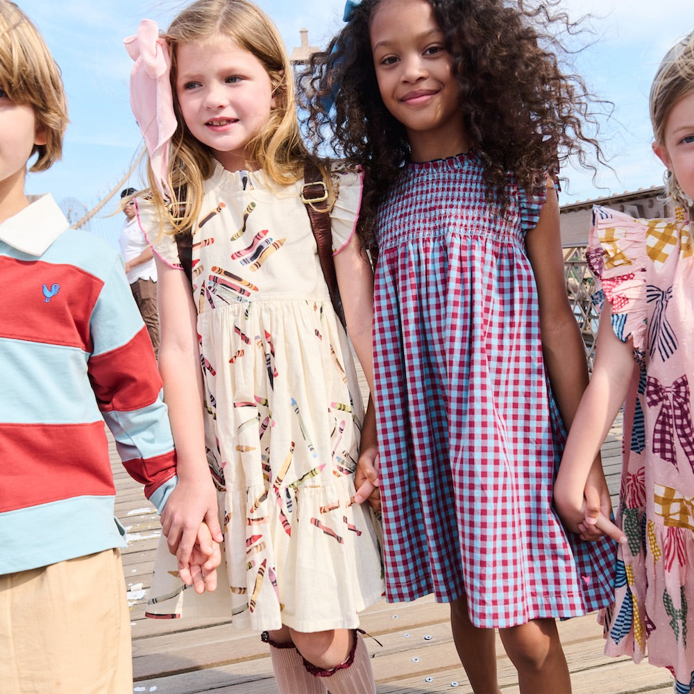 Four children stand holding hands outdoors, dressed in colorful clothes; two girls are in the center, one wearing a patterned cream Girls’ Stevie Dress by Pink Chicken and the other in a vibrant red & blue check plaid dress.