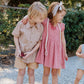 Three young children stand outdoors: a boy in a Pink Chicken Boys’ Jack Shirt, a girl in matching Cocoa Gingham outfits with her arm around him, and a toddler in a blue romper close by.