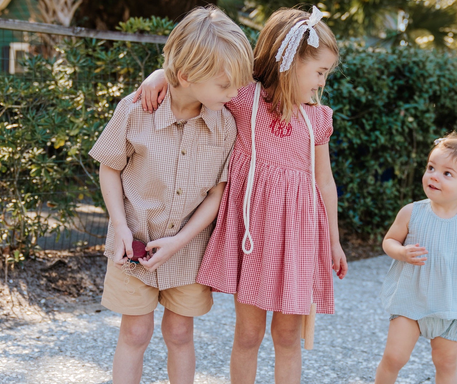 Three young children stand outdoors: a boy in a Pink Chicken Boys’ Jack Shirt, a girl in matching Cocoa Gingham outfits with her arm around him, and a toddler in a blue romper close by.