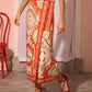 A person in a patterned red and beige Paloma Wrap Shirt by Hunter Bell NYC, a sleeveless polka dot top, and red lace-up flats stands indoors near a red chair.