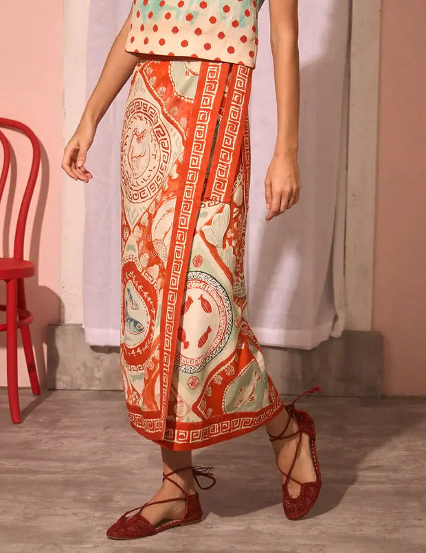 A person in a patterned red and beige Paloma Wrap Shirt by Hunter Bell NYC, a sleeveless polka dot top, and red lace-up flats stands indoors near a red chair.