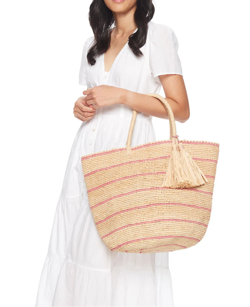 A woman in a white short-sleeve dress holds the Mar Y Sol Costa Tote, a spacious and stylish bag woven with pink stripes and adorned with a decorative tassel.