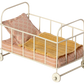 The Maileg Cot Bed, Micro - Rose by Maileg is a small, white, metal doll crib with four wheels. It comes complete with a pink and white striped mattress designed for small mice, along with a yellow blanket and a pink patterned pillow.