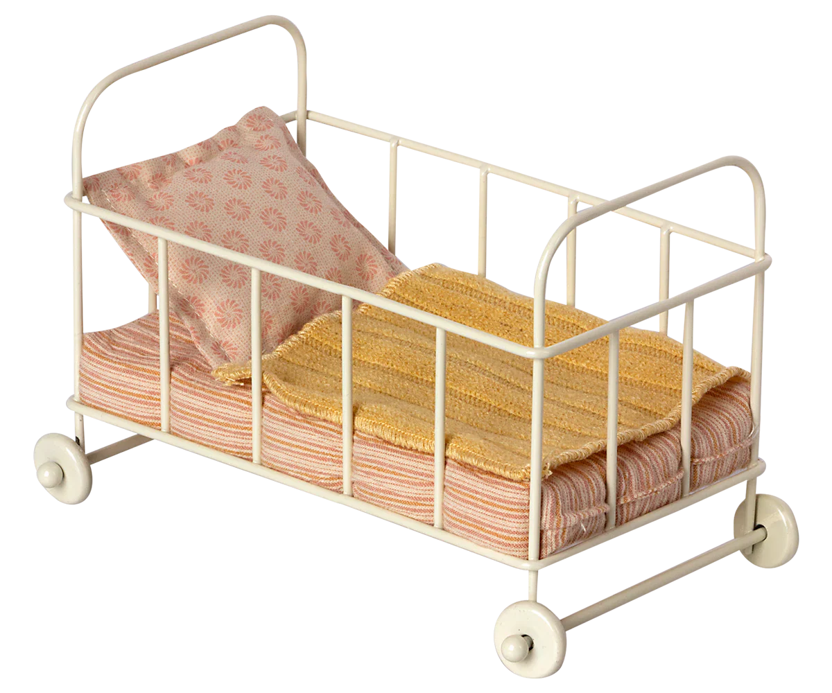 The Maileg Cot Bed, Micro - Rose by Maileg is a small, white, metal doll crib with four wheels. It comes complete with a pink and white striped mattress designed for small mice, along with a yellow blanket and a pink patterned pillow.