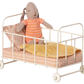 A Maileg Cot Bed, Micro - Rose, by Maileg features a stuffed rabbit dressed in an orange-striped outfit sitting inside the white metal frame, complete with an orange-striped mattress, a pink pillow, and a yellow blanket.