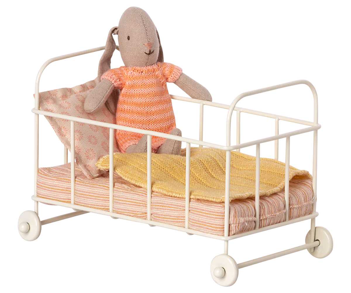 A Maileg Cot Bed, Micro - Rose, by Maileg features a stuffed rabbit dressed in an orange-striped outfit sitting inside the white metal frame, complete with an orange-striped mattress, a pink pillow, and a yellow blanket.