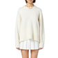 A woman wears the Guest in Residence Cozy Crew Cashmere Sweater—an oversized cream knit with ribbed neck trim and dropped shoulders—styled with a short white pleated skirt and chunky necklace, against a plain white background.