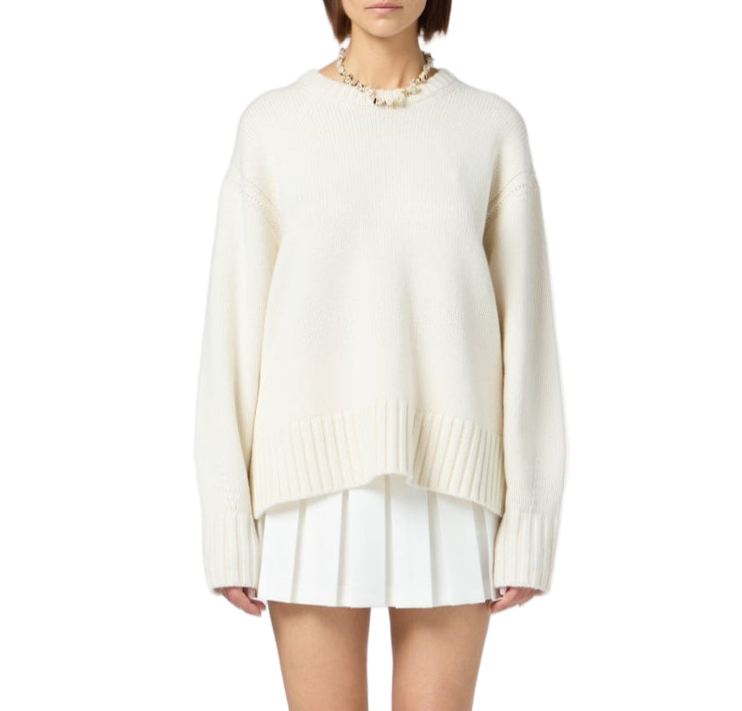 A woman wears the Guest in Residence Cozy Crew Cashmere Sweater—an oversized cream knit with ribbed neck trim and dropped shoulders—styled with a short white pleated skirt and chunky necklace, against a plain white background.