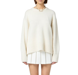 A woman wears the Guest in Residence Cozy Crew Cashmere Sweater—an oversized cream knit with ribbed neck trim and dropped shoulders—styled with a short white pleated skirt and chunky necklace, against a plain white background.