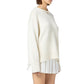 A woman wears the Guest in Residence Cozy Crew Cashmere Sweater in cream, featuring a ribbed neck trim and dropped shoulders, paired with a white pleated skirt against a plain white background.