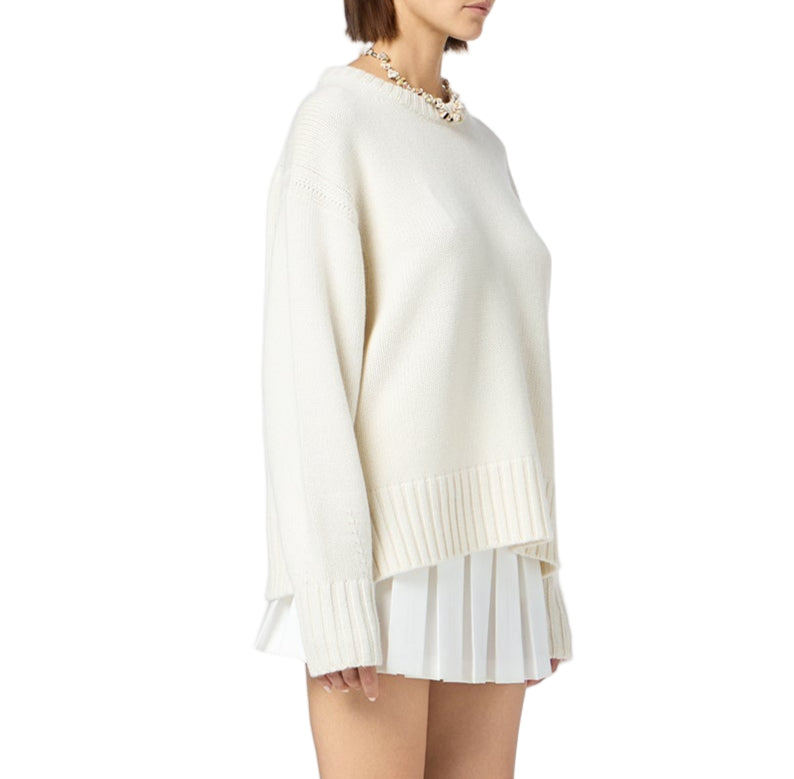 A woman wears the Guest in Residence Cozy Crew Cashmere Sweater in cream, featuring a ribbed neck trim and dropped shoulders, paired with a white pleated skirt against a plain white background.