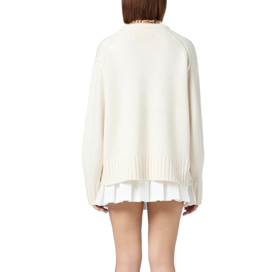 A person stands with their back to the camera, wearing the Guest in Residence Cozy Crew Cashmere Sweater—an oversized white knit with a ribbed neck trim—paired with a short white pleated skirt, set against a plain white background.