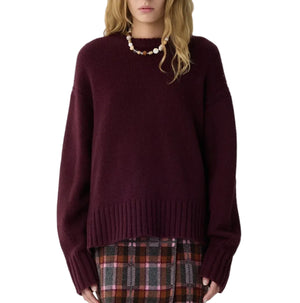 A person wearing the Guest in Residence Cozy Crew Cashmere Sweater in dark maroon, paired with a beaded necklace and a brown plaid skirt, stands against a white background.