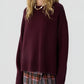A woman wearing the Guest in Residence Cozy Crew Cashmere Sweater in an oversized maroon fit, paired with a plaid mini skirt featuring a front slit and a beaded necklace, stands against a plain light background.