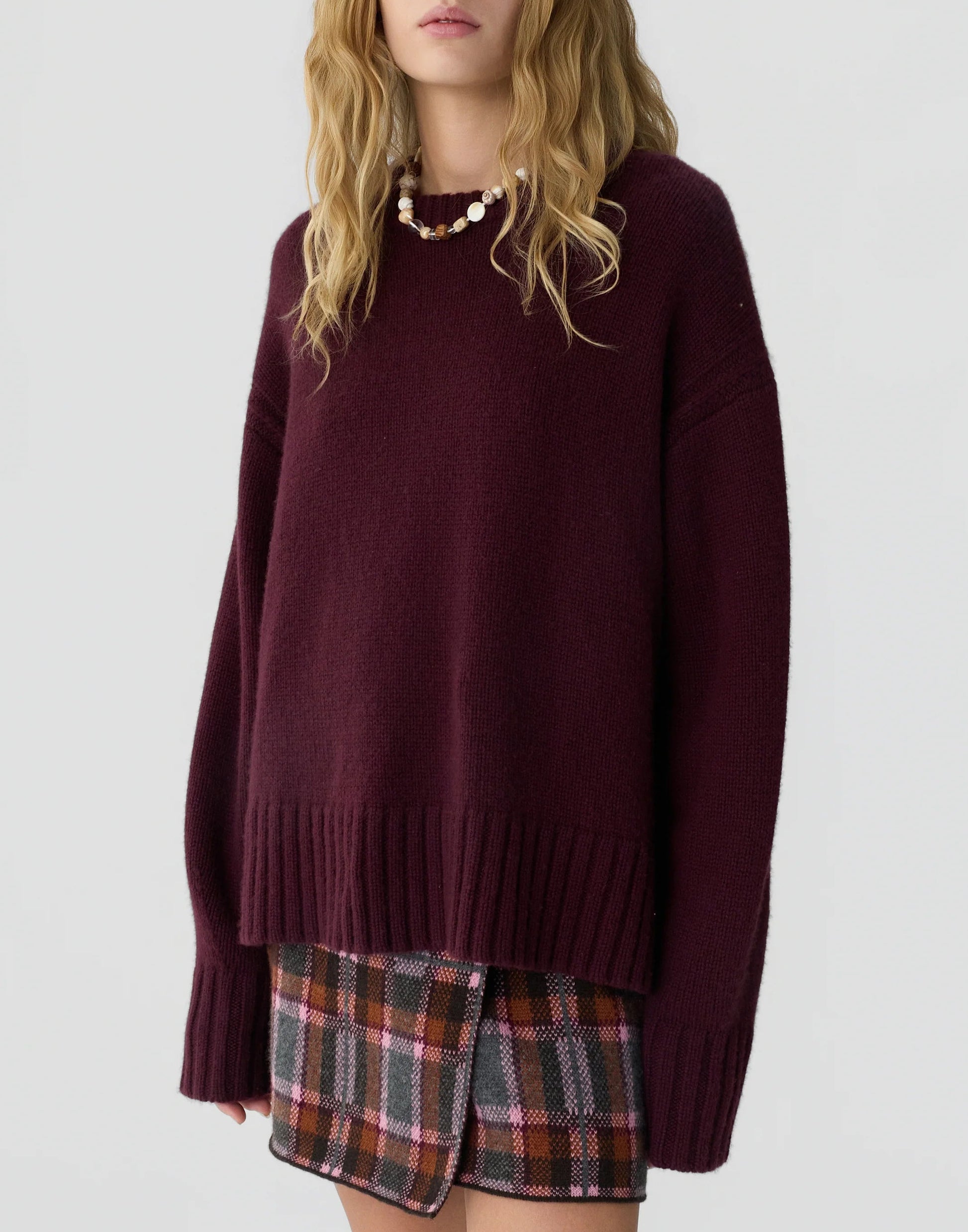 A woman wearing the Guest in Residence Cozy Crew Cashmere Sweater in an oversized maroon fit, paired with a plaid mini skirt featuring a front slit and a beaded necklace, stands against a plain light background.