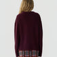 A person with long, wavy blonde hair is shown from the back against a plain light background, wearing the Guest in Residence Cozy Crew Cashmere Sweater in burgundy paired with a plaid skirt.