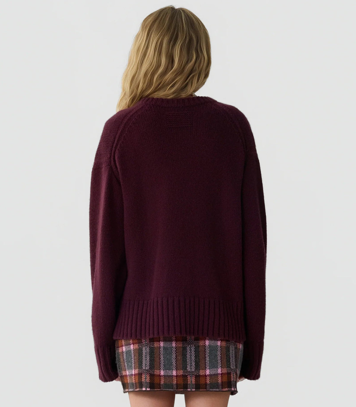 A person with long, wavy blonde hair is shown from the back against a plain light background, wearing the Guest in Residence Cozy Crew Cashmere Sweater in burgundy paired with a plaid skirt.