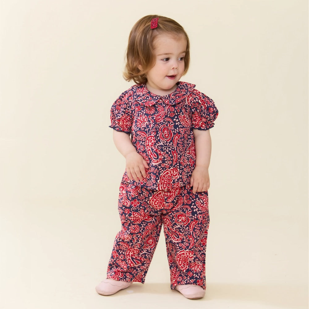A young child with light brown hair and a pink hairclip wears the Pink Chicken Baby Girls’ Dawson 2-Piece Set, featuring a red and blue paisley vines pattern, paired with pink shoes and standing against a plain light background.