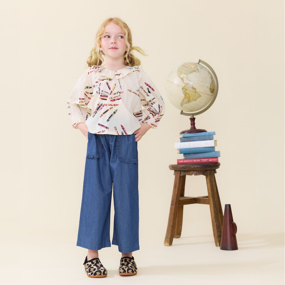 A young girl stands with her hands in the pockets of her Pink Chicken Girls’ Bailey Ruffle Pant, wearing a patterned shirt and blue pants, next to a stool with stacked books, a globe, and a cone-shaped object on the floor.
