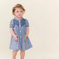 A young child with light brown hair and a bow stands wearing the Pink Chicken Baby Girls’ Lucia Dress Set—a blue-patterned, short-sleeve dress featuring delicate diamond smocking that showcases timeless heritage craftsmanship—looking slightly upward against a plain light background.