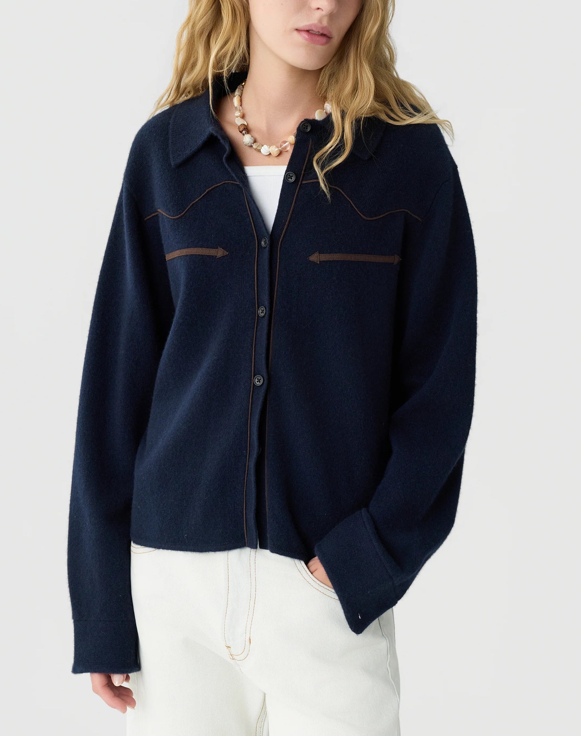 A person with long blonde hair wears the Guest in Residence Cropped Western Shirt in navy with brown stitching, styled over a white top and paired with white pants and a beaded necklace, set against a plain background.