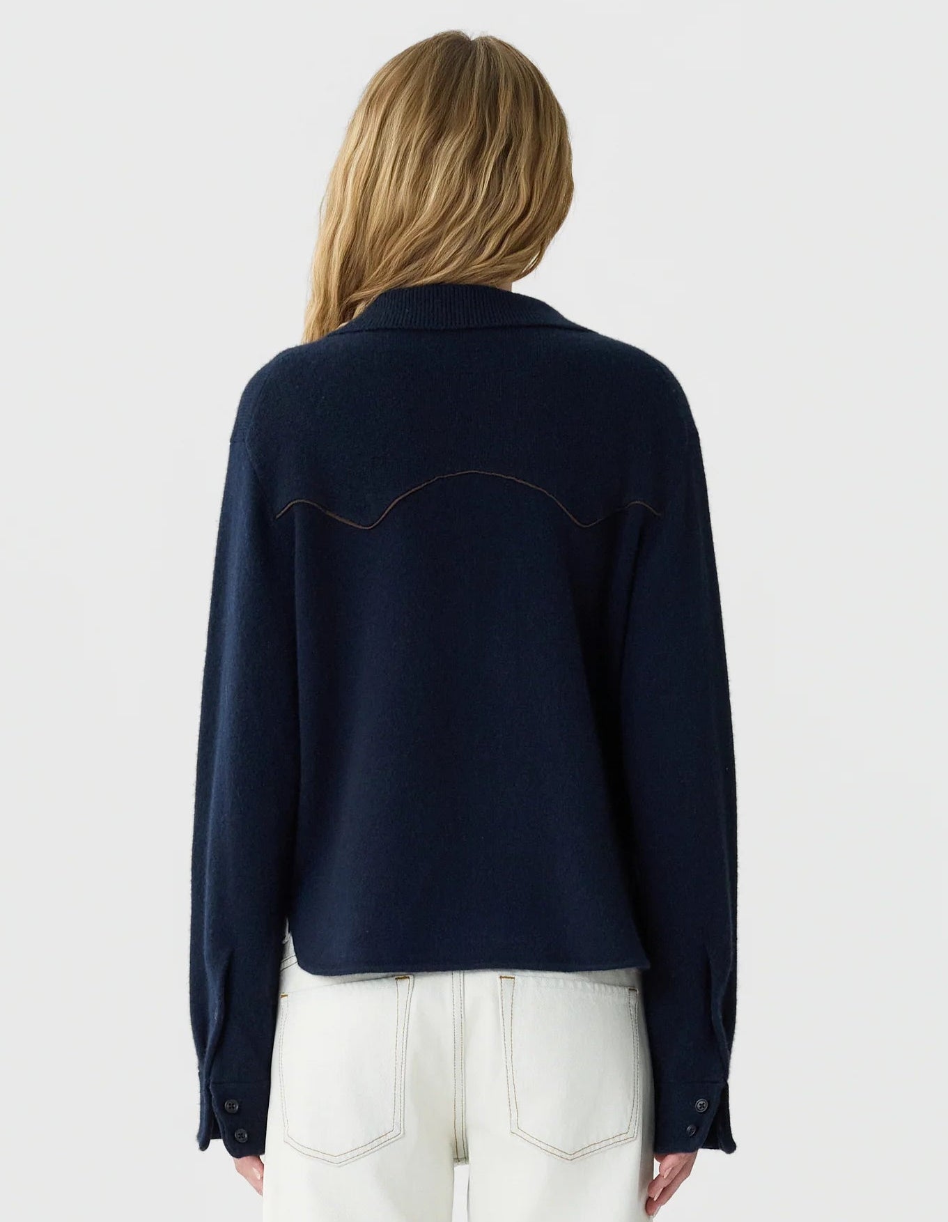 A person with long, wavy blonde hair wears the Guest in Residence Cropped Western Shirt in dark navy, styled with off-white jeans, standing facing away from the camera against a plain light background.