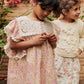 Two young girls in matching Girls' Marise Dresses by Louise Misha stand outside, one looking at her hands and the other smiling. Red flowers and greenery beautifully enhance their cotton voile outfits.