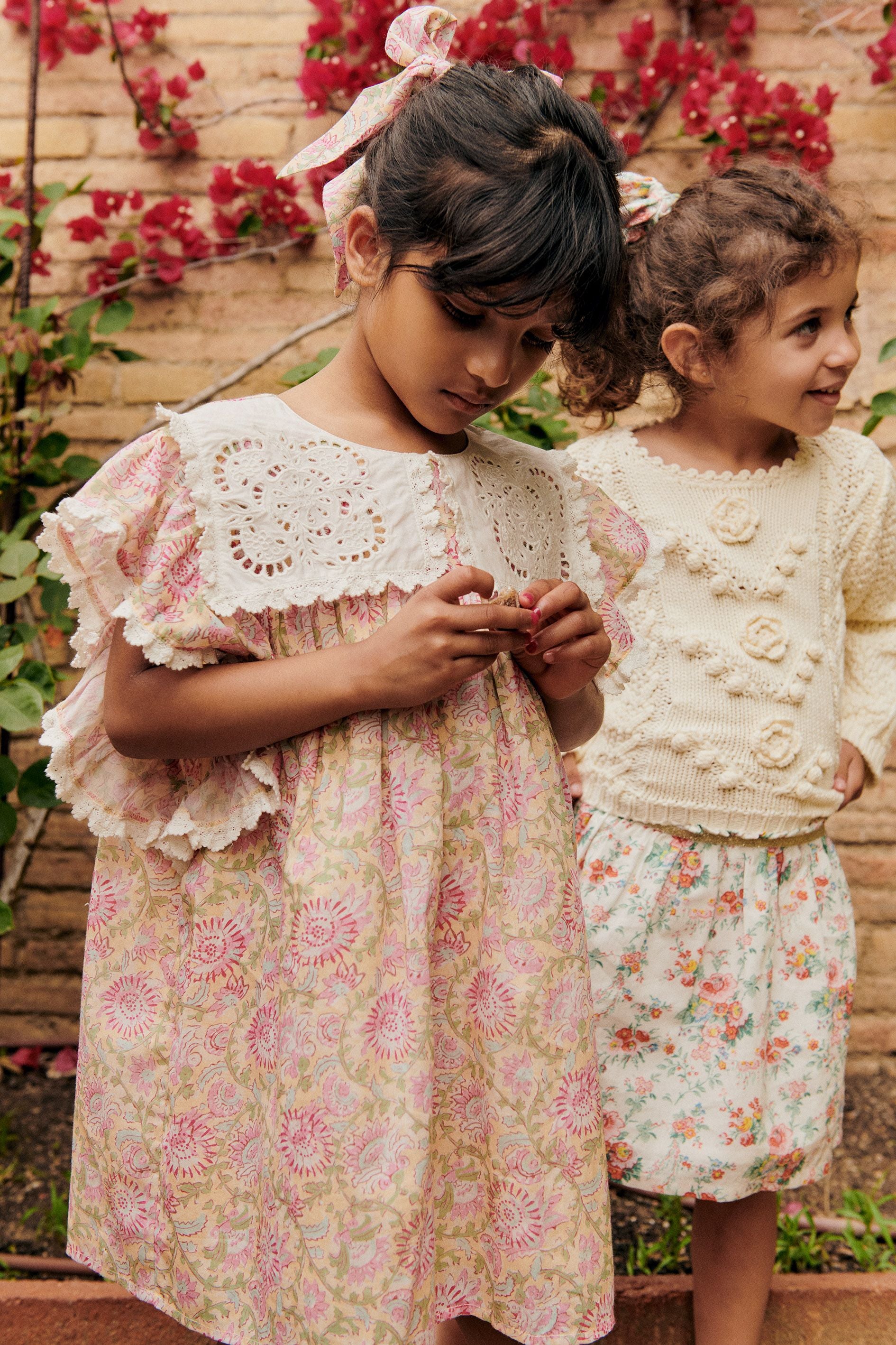 Two young girls in matching Girls' Marise Dresses by Louise Misha stand outside, one looking at her hands and the other smiling. Red flowers and greenery beautifully enhance their cotton voile outfits.