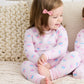 A young child with light brown hair and a pink bow sits on a bed, wearing the Kissy Kissy Baby Girls' and Girls' Cupcake Party Pajama Set.