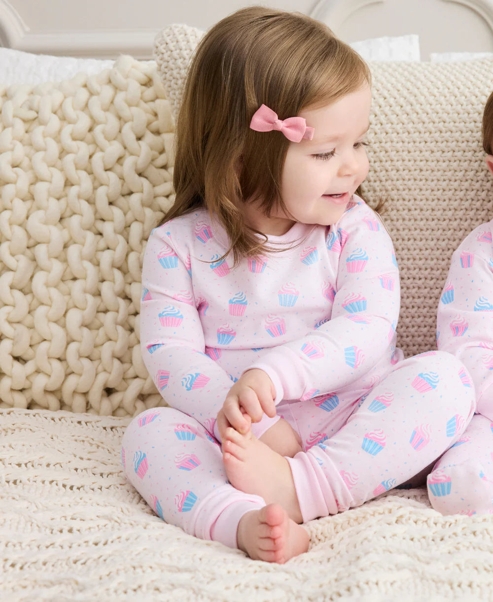 A young child with light brown hair and a pink bow sits on a bed, wearing the Kissy Kissy Baby Girls' and Girls' Cupcake Party Pajama Set.