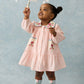 A young girl wearing the Louise Misha Ysee Dress stands on a light background, reaching up to touch white hanging decorations.