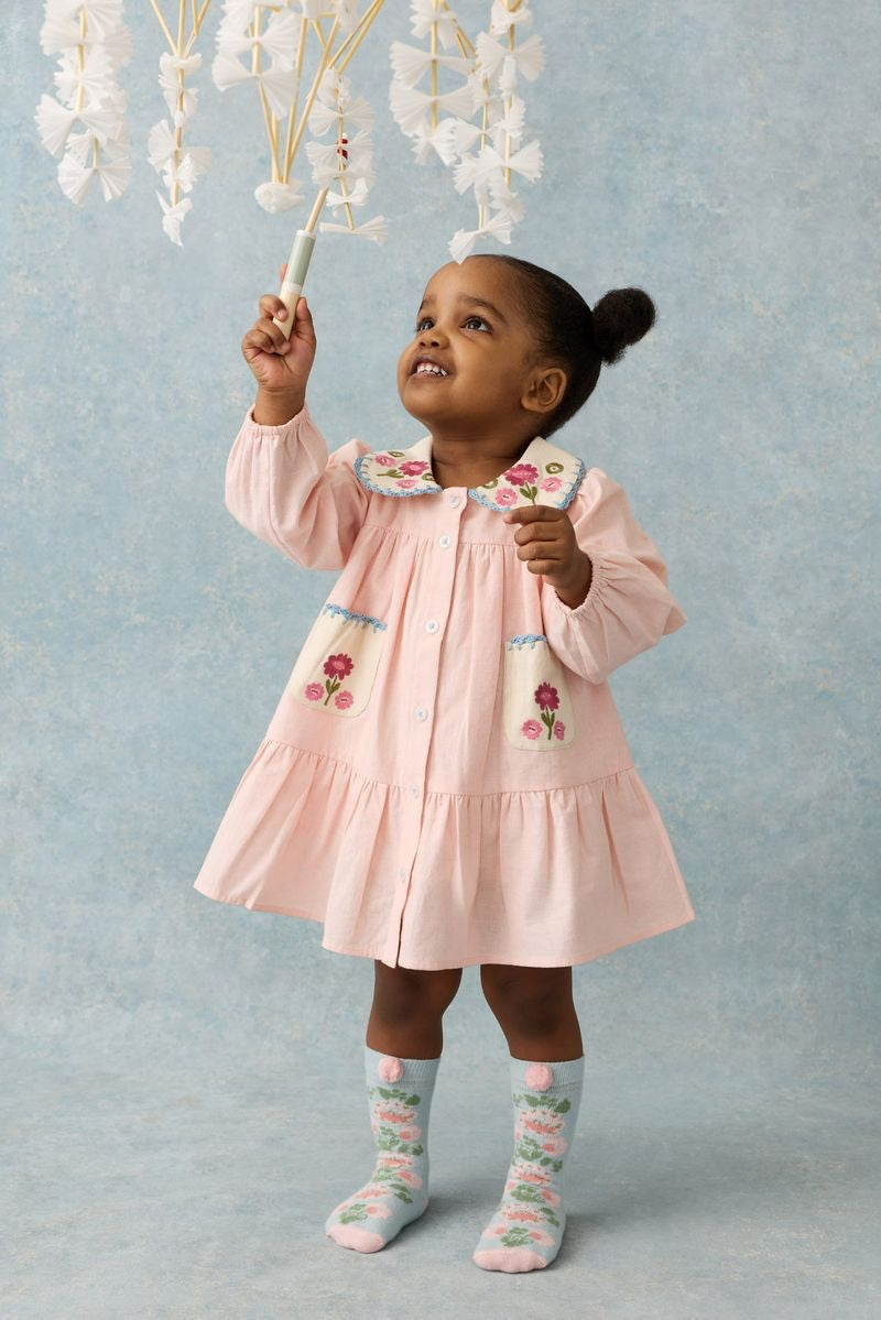 A young girl wearing the Louise Misha Ysee Dress stands on a light background, reaching up to touch white hanging decorations.