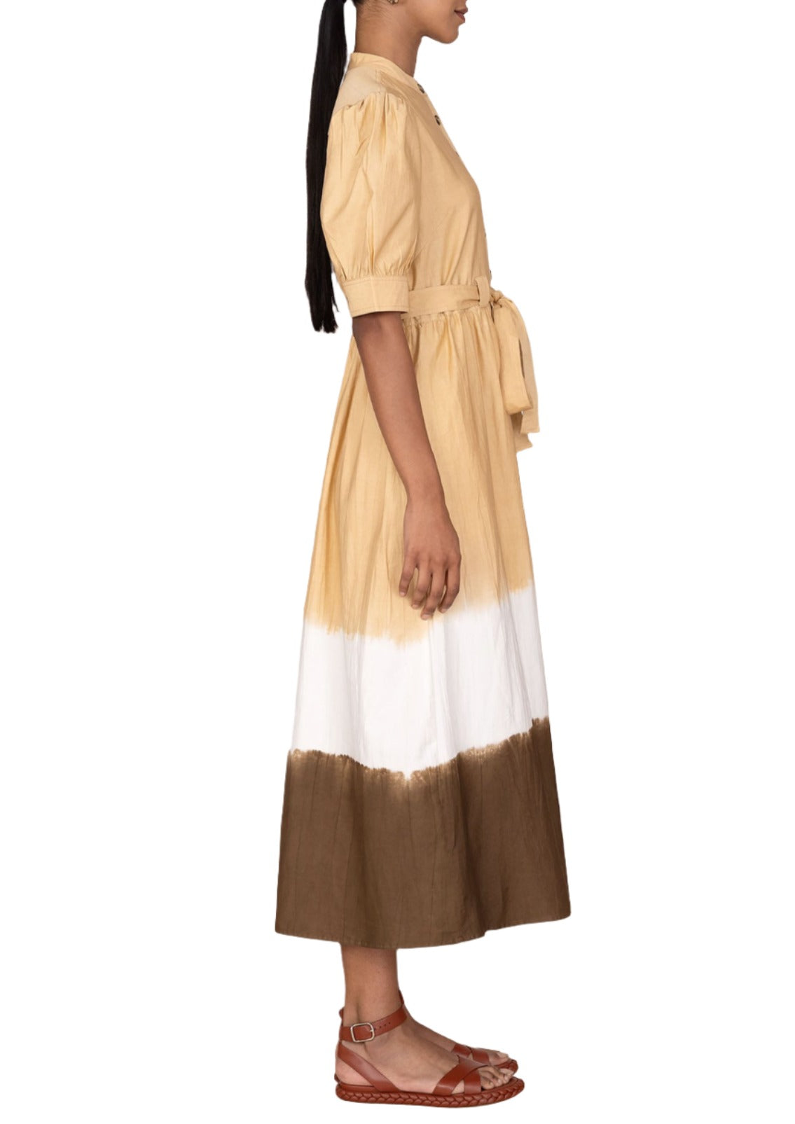 A woman stands facing left wearing a Banjanan Daffodil Dress in beige, white, and brown with short puff sleeves and a waist tie. She is also wearing brown, strappy flat sandals.