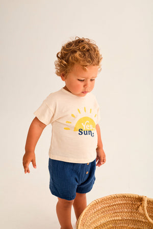A young child with curly hair wears Cozmo's Baby Boys' Dale Shorts and a "Yes, Sun" t-shirt, standing by a woven basket against a plain light background.