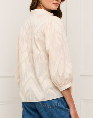 A woman wearing the Figue Danilia Top—a light-patterned band collar blouse with three-quarter sleeves—paired with blue jeans, stands in front of a white wall, facing slightly away from the camera.
