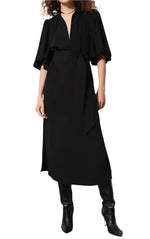 A person styled in the Figue Dani Dress, showcasing a '70s chic look. The black dress from Figue features short puff sleeves, a wrap belt, and a fashionable V-neckline, complemented by knee-high black boots.