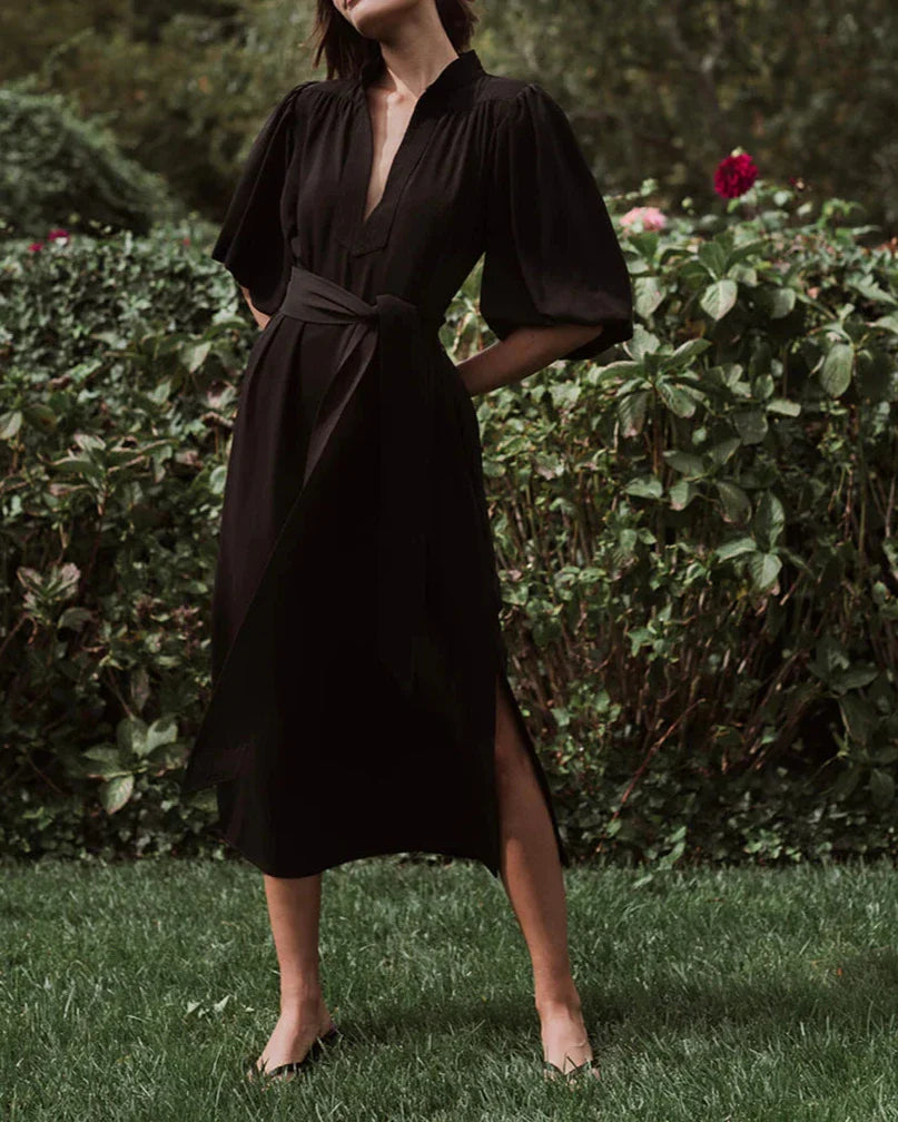 A person stands on grass, wearing the sophisticated Figue Dani Dress with puffed sleeves and a tied waist, radiating '70s chic style amid lush green foliage and pink flowers.