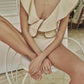 Wearing the Johanna Ortiz Crinkle Beaches One Piece in cream, a person sits on a white chair with arms and legs crossed, accessorized with a black necklace featuring a gold wishbone pendant—a chic swimwear statement.