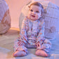 A baby in the Baby Girls’ Arabella Ruffle Romper by Magnetic Me sits on a soft surface, smiling in front of two textured round cushions.